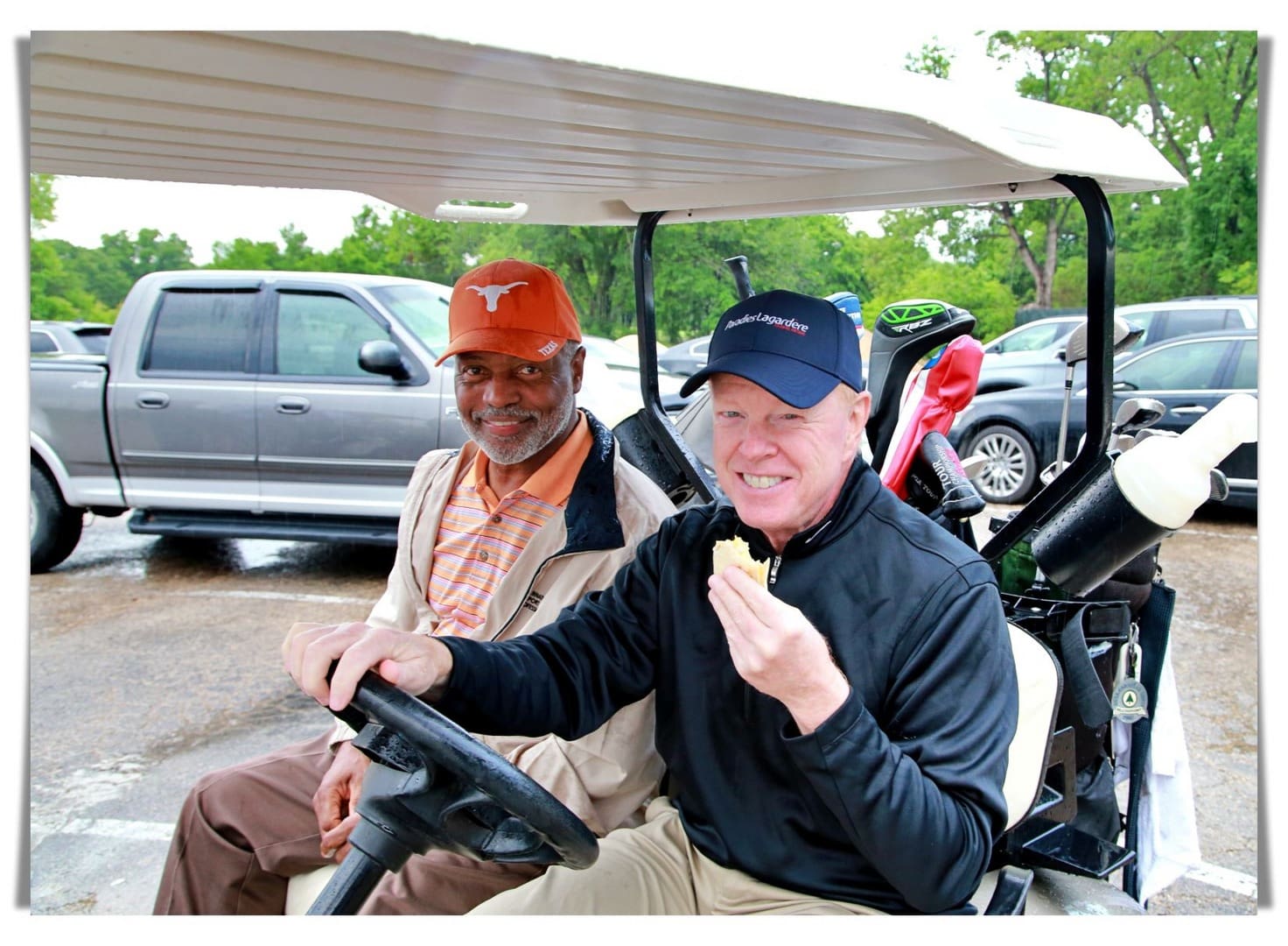 Two men sitting in a golf cart eating food.