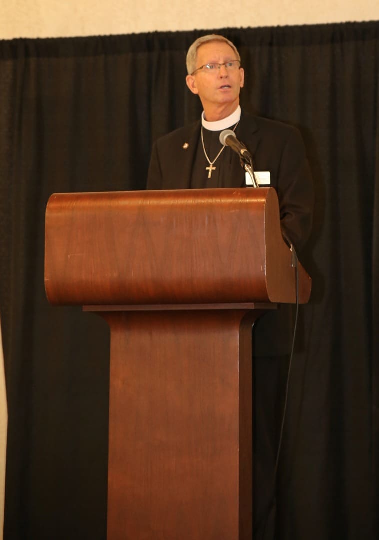 A priest standing at a podium in front of a black background.